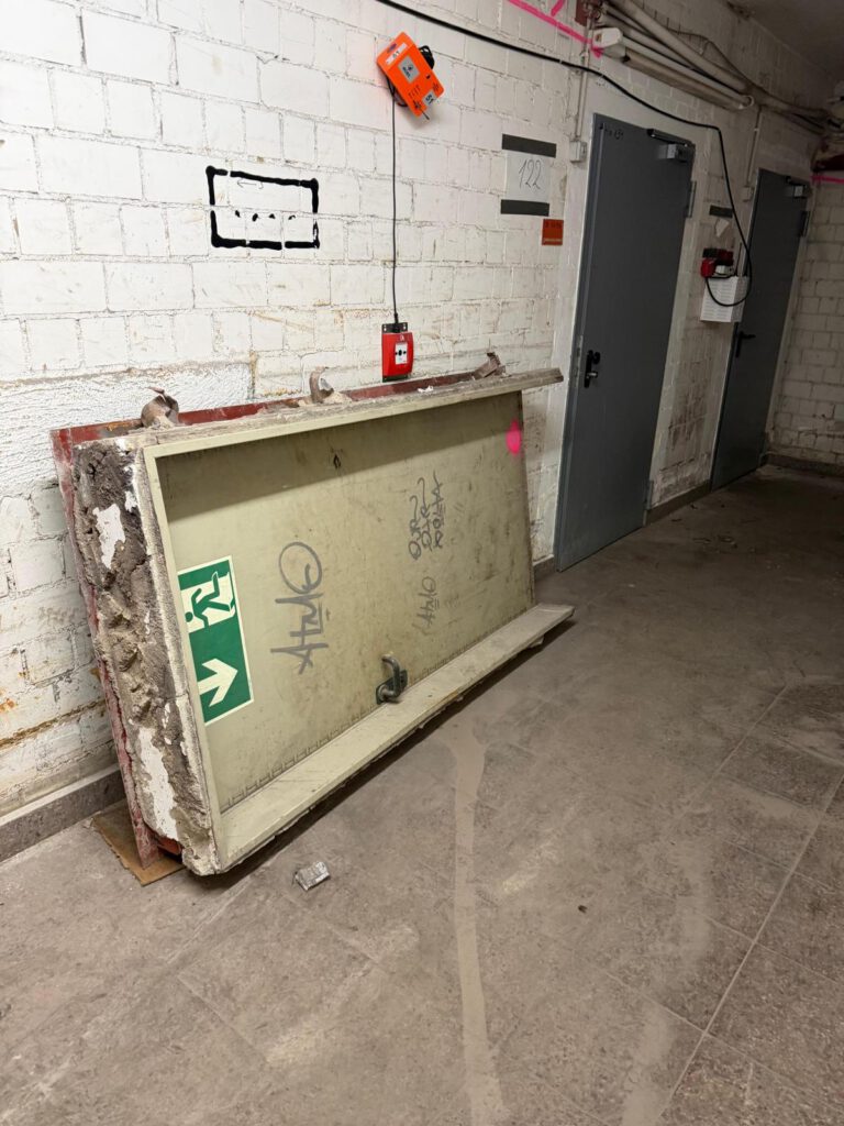 Asbestplatte lehnt an einer weißen Wand in einem dunklen Kellerflur