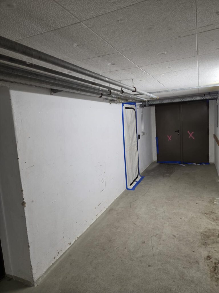 Kellerflur mit Rohrleitungen, blau abgeklebter Folientür und markierten Doppeltüren.